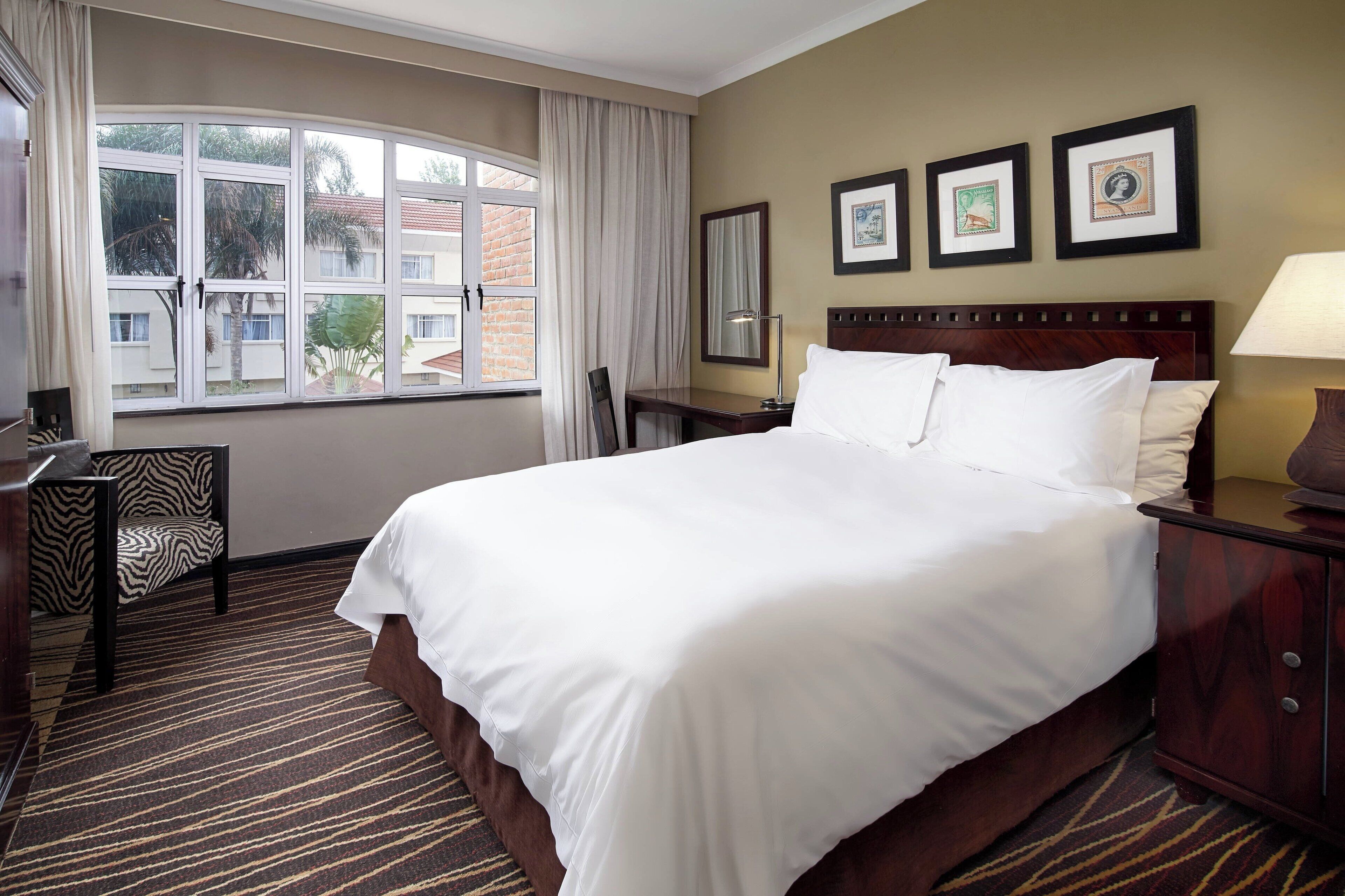 A hotel room with a large bed, a desk, a chair, a window with curtains, and framed pictures on the wall.