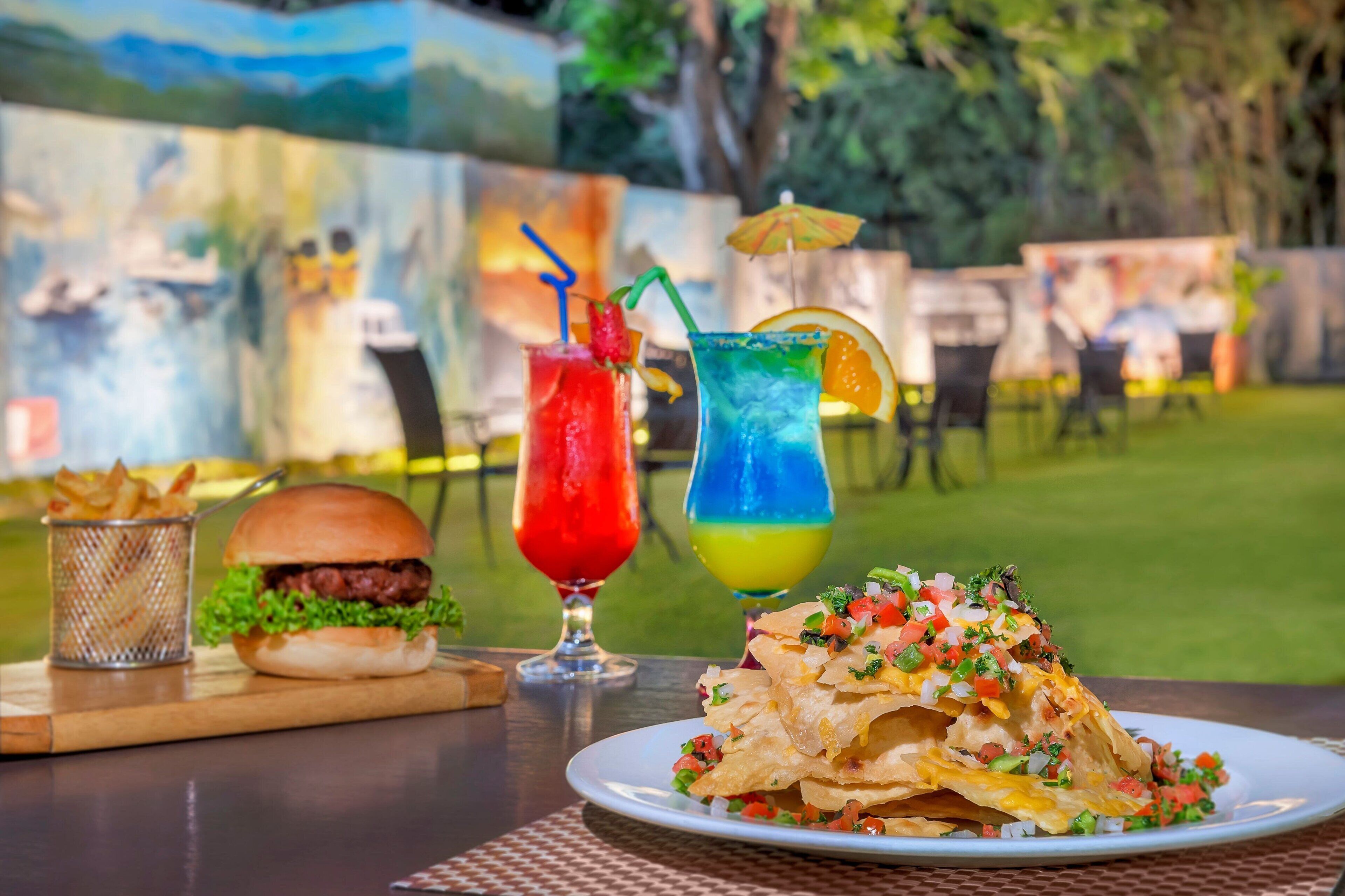 Comida con nachos, una hamburguesa y dos bebidas sobre una mesa al aire libre.