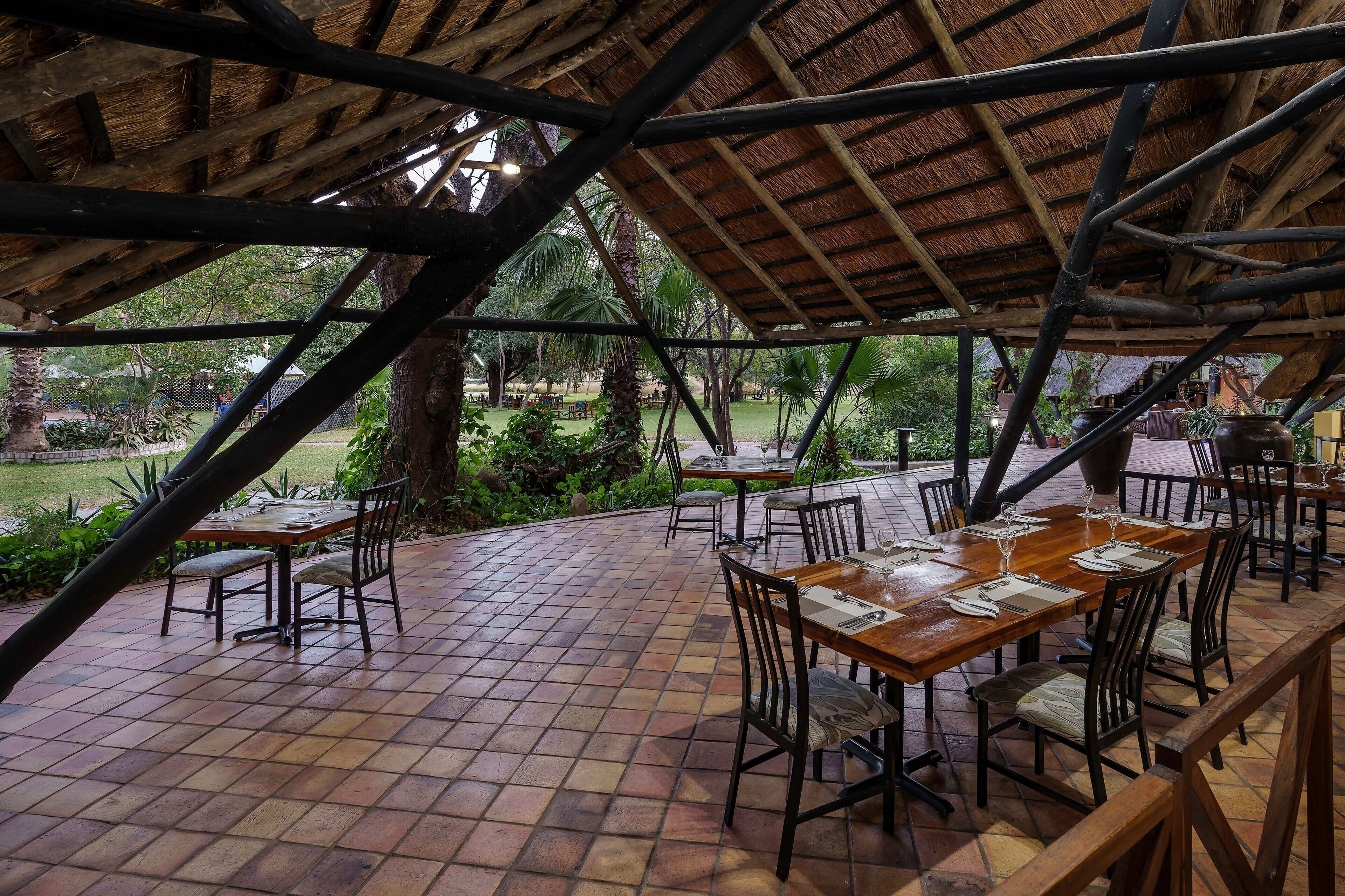 An outdoor dining area with wooden tables and chairs, a tiled floor, and a thatched roof structure.