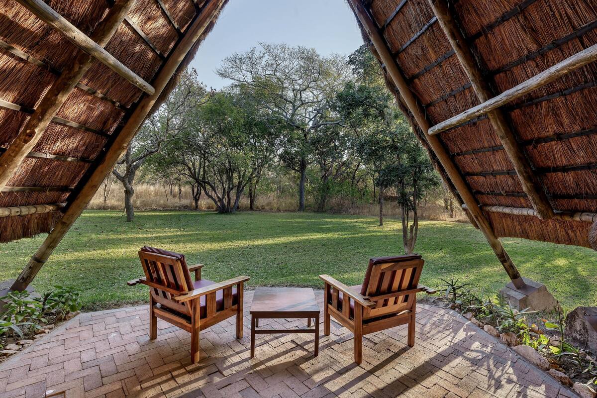 An outdoor seating area with two wooden chairs and a table, surrounded by a brick patio and a view of a grassy landscape with trees.