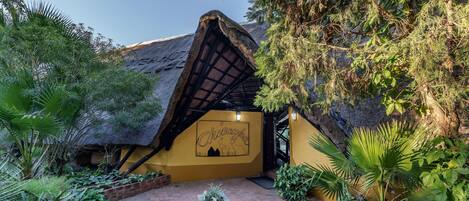 A thatched-roof hut with a mural, surrounded by lush greenery and a brick pathway.