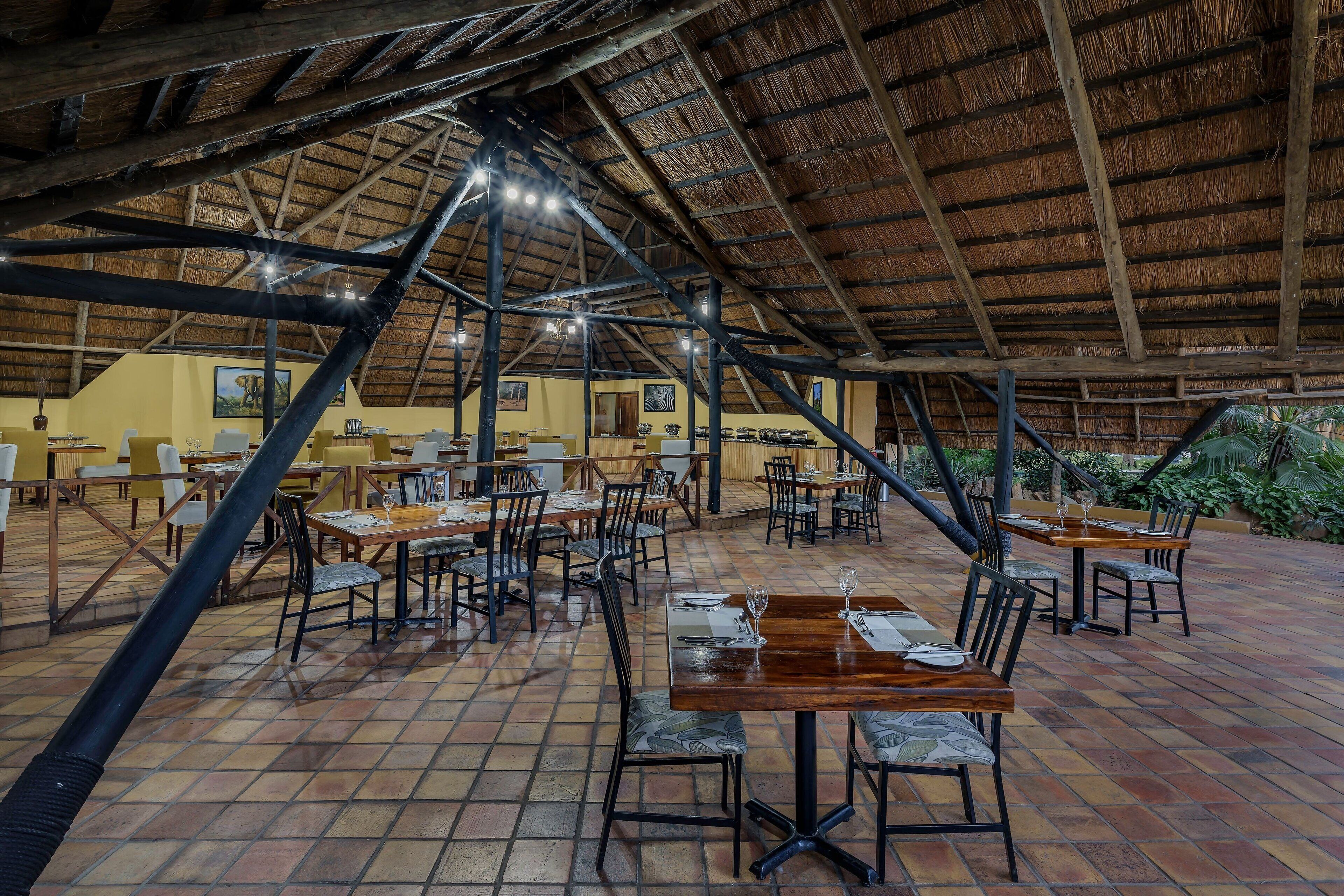 An indoor dining area with wooden tables and chairs, a tiled floor, and a thatched roof structure.