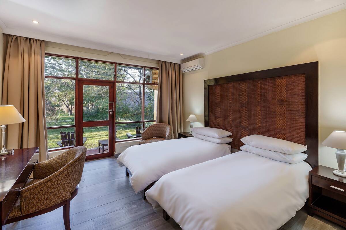 A hotel room with two beds, a desk, a chair, a window with curtains, and an air conditioning unit.