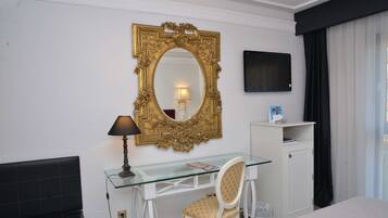 Habitación de hotel con un escritorio de vidrio, una silla blanca, un espejo con marco dorado, una televisión y una cama.