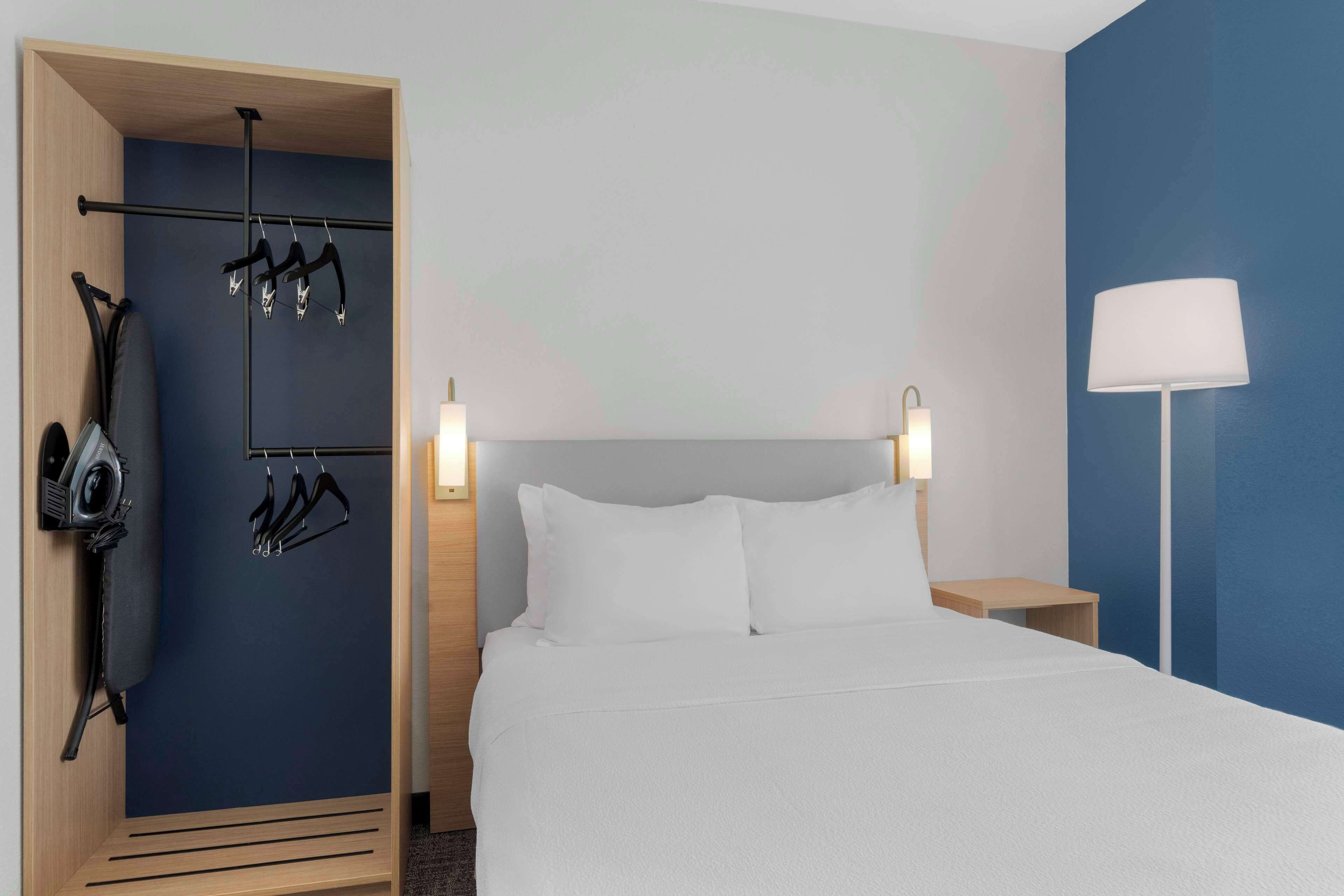 A hotel room with a bed, a wardrobe with hanging clothes, a lamp, and a nightstand.