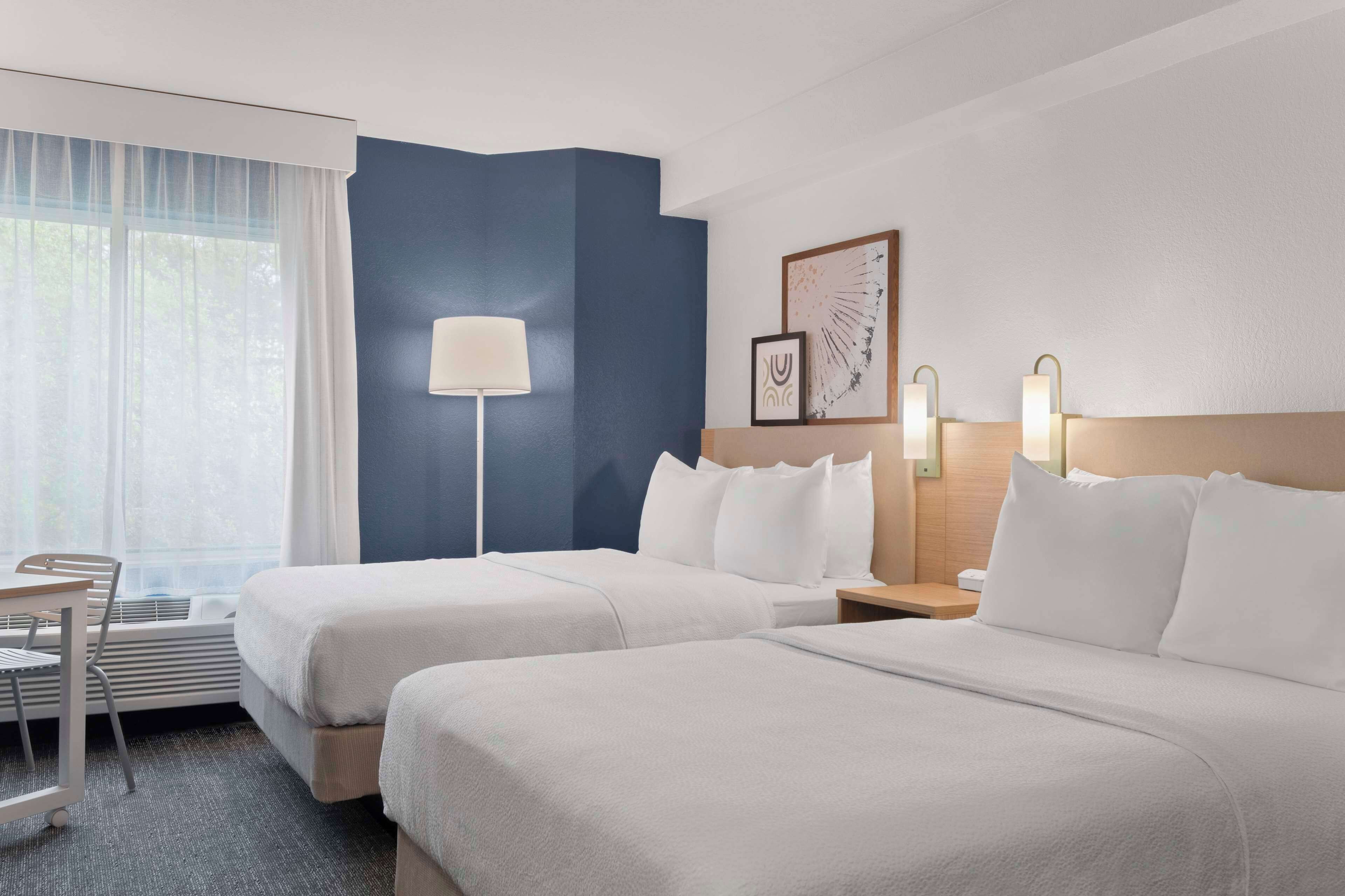 Une chambre d’hôtel avec deux lits, un bureau, une chaise, une lampe et une œuvre d’art encadrée au mur.