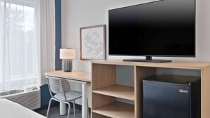 Habitació d'hotel amb llit, escriptori i cadira, televisió, nevera i finestra amb cortines.