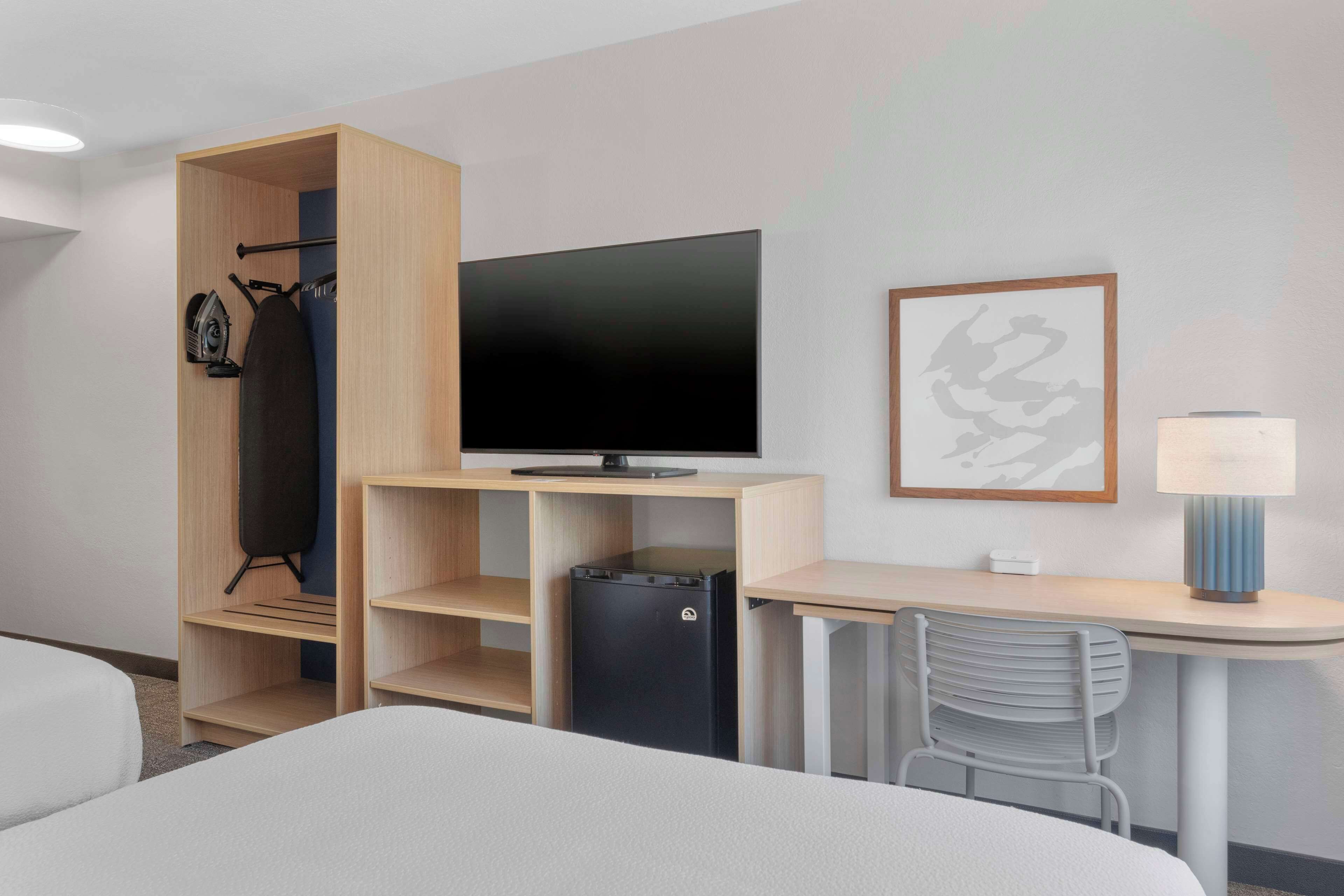 A hotel room with a bed, a desk with a chair, a television, a small refrigerator, and a wardrobe with hanging clothes.