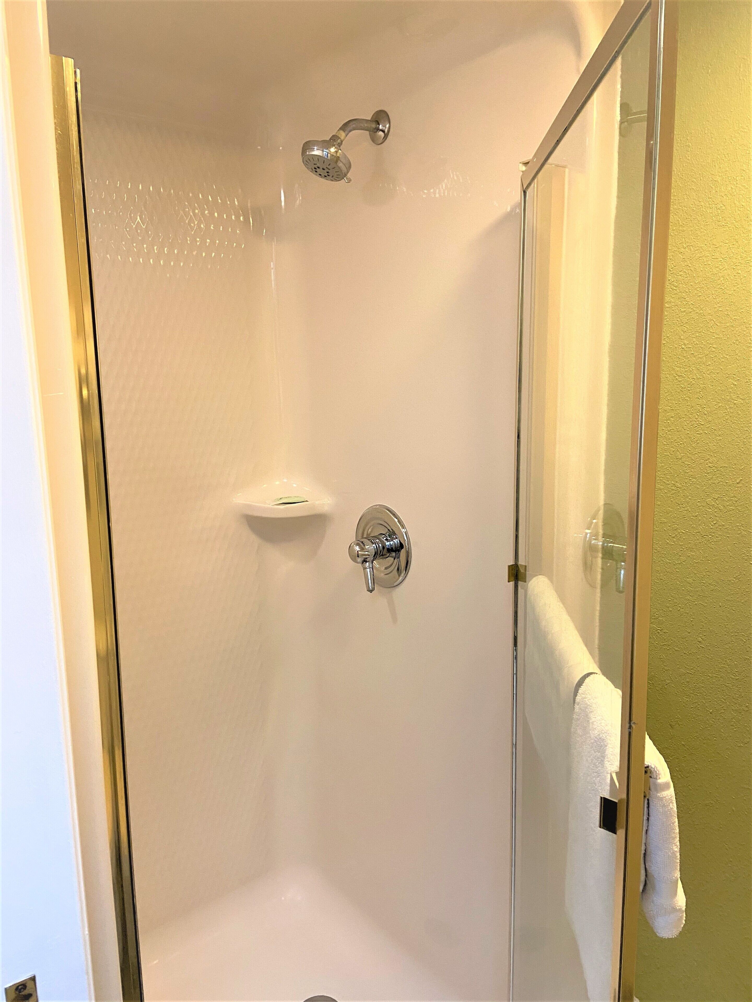 A walk-in shower with a glass door, a handheld showerhead, a wall-mounted faucet, and a small shelf.