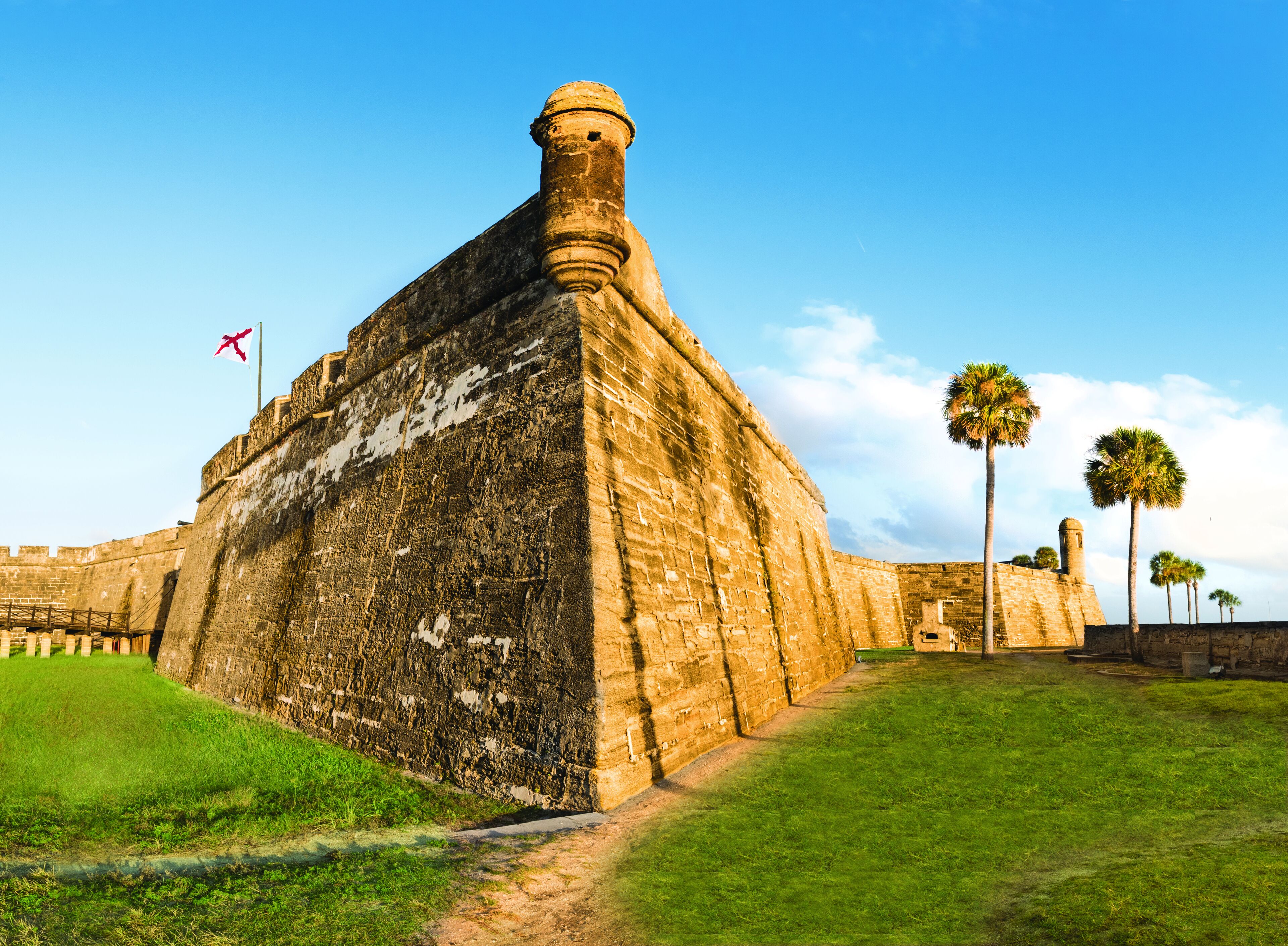 Una fortaleza histórica con una bandera, palmeras y un cielo despejado.
