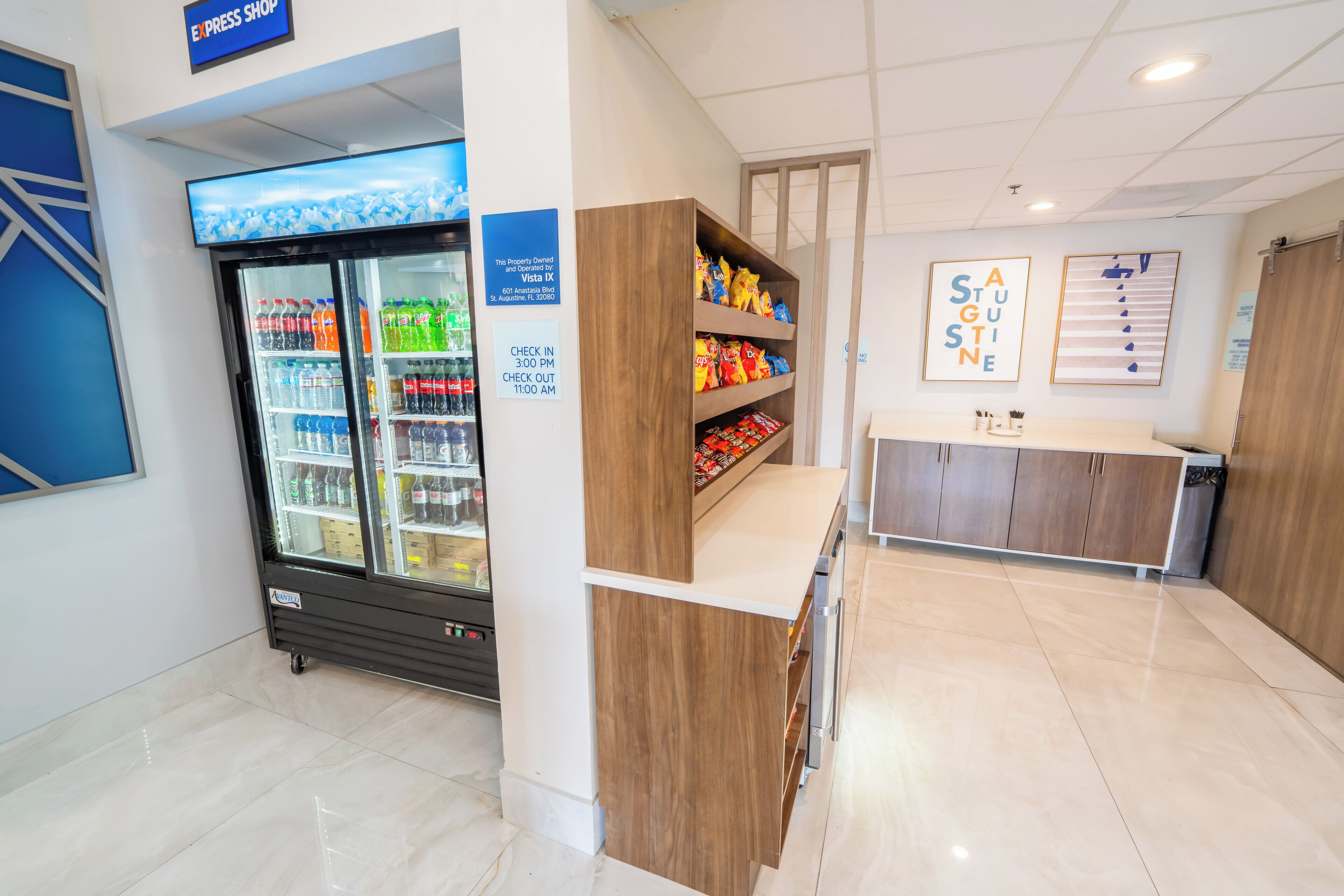Un interior limpio y moderno con un refrigerador de autoservicio lleno de diversas bebidas, una encimera de madera con aperitivos y una zona de recepción con un cartel que indica los horarios de registro de entrada y salida.