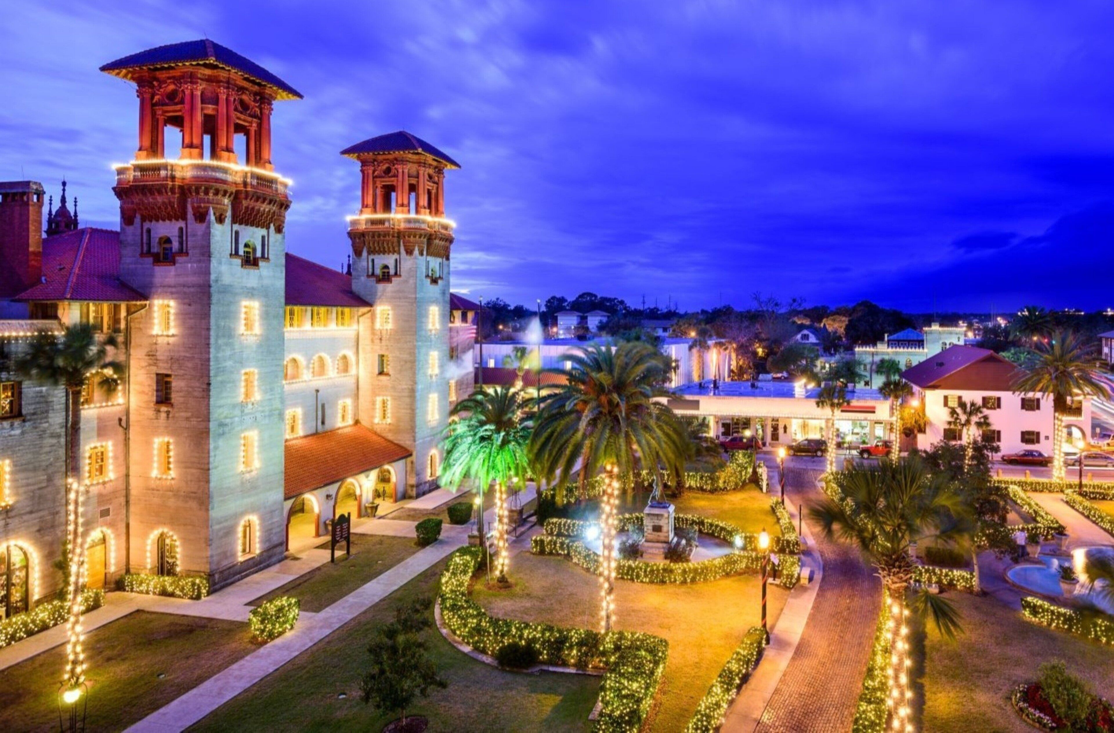 A historic building with two towers, a courtyard with palm trees, and a well-lit pathway.