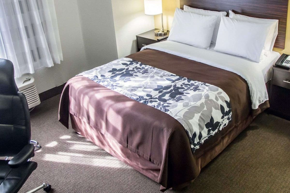 Habitación de hotel con cama, mesita de noche, lámpara, silla y ventana con cortinas.