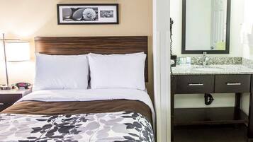 Habitación de hotel con cama, mesita de noche, lámpara, una colección de fotos enmarcadas en la pared y baño con lavamanos y espejo.