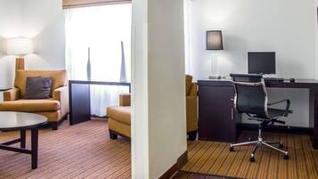 A hotel room with a sofa, a desk with a computer, a lamp, and a window with blinds.