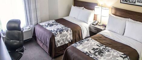 A hotel room with two beds, a desk, and a chair. There are framed pictures on the wall and a lamp on the nightstand.