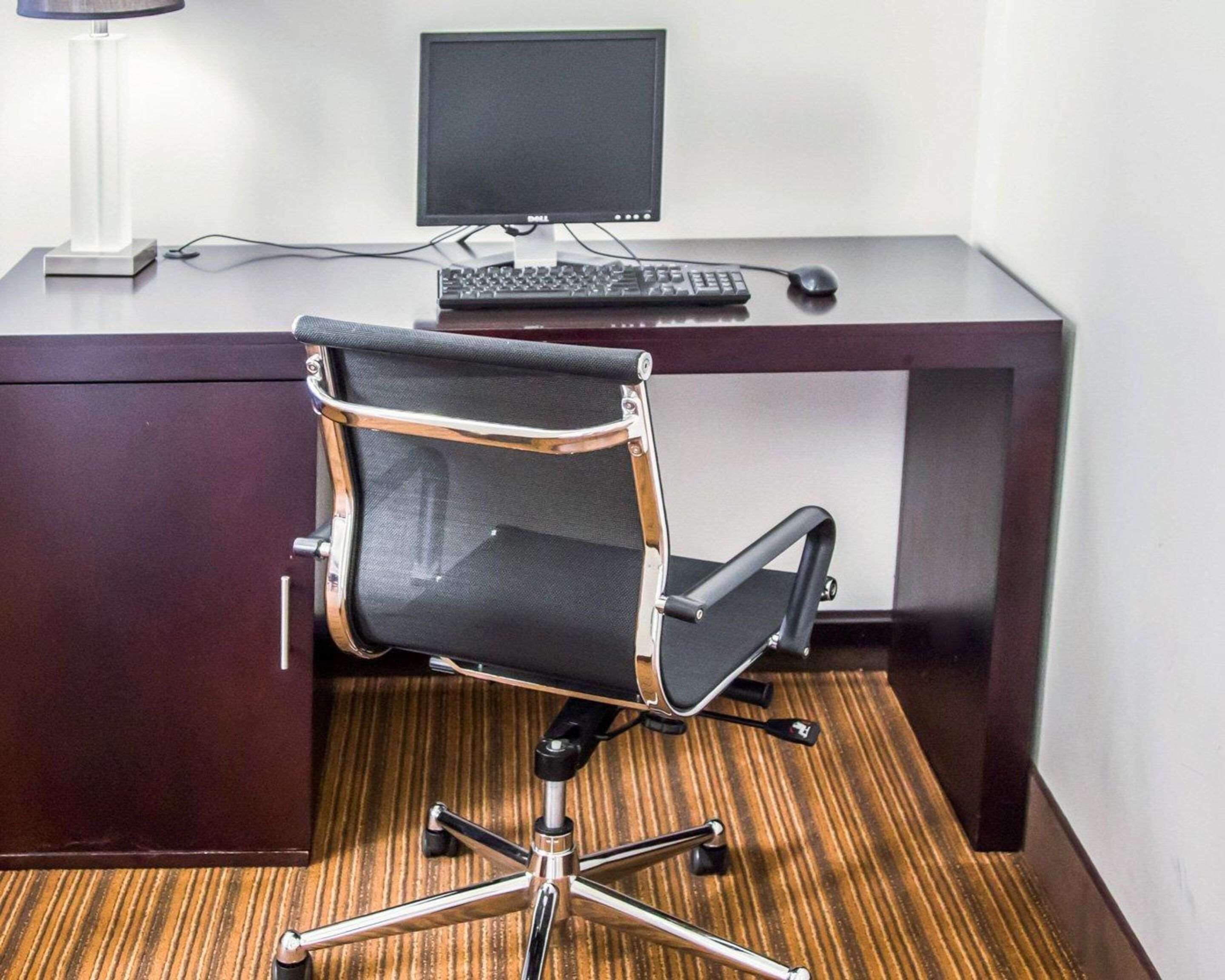 A modern office chair with a mesh backrest and metal frame, positioned in front of a dark wooden desk with a computer monitor, keyboard, and mouse.
