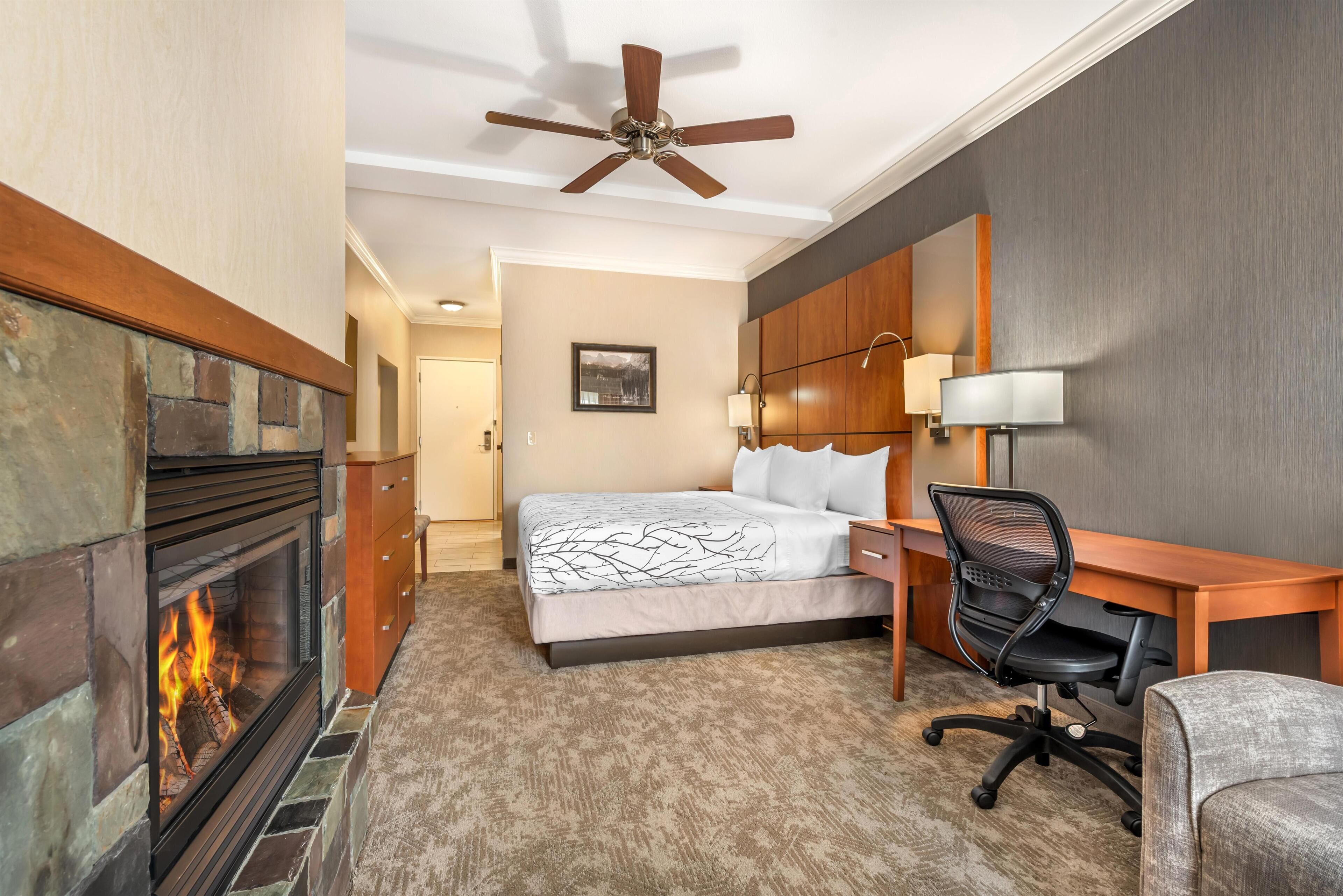 A hotel room with a fireplace, a bed, a desk, a chair, and a ceiling fan.