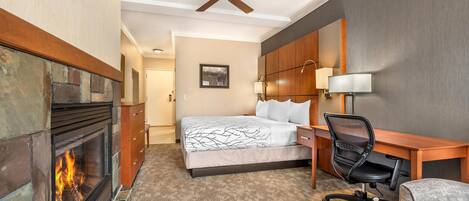 A hotel room with a fireplace, a bed, a desk, a chair, and a ceiling fan.