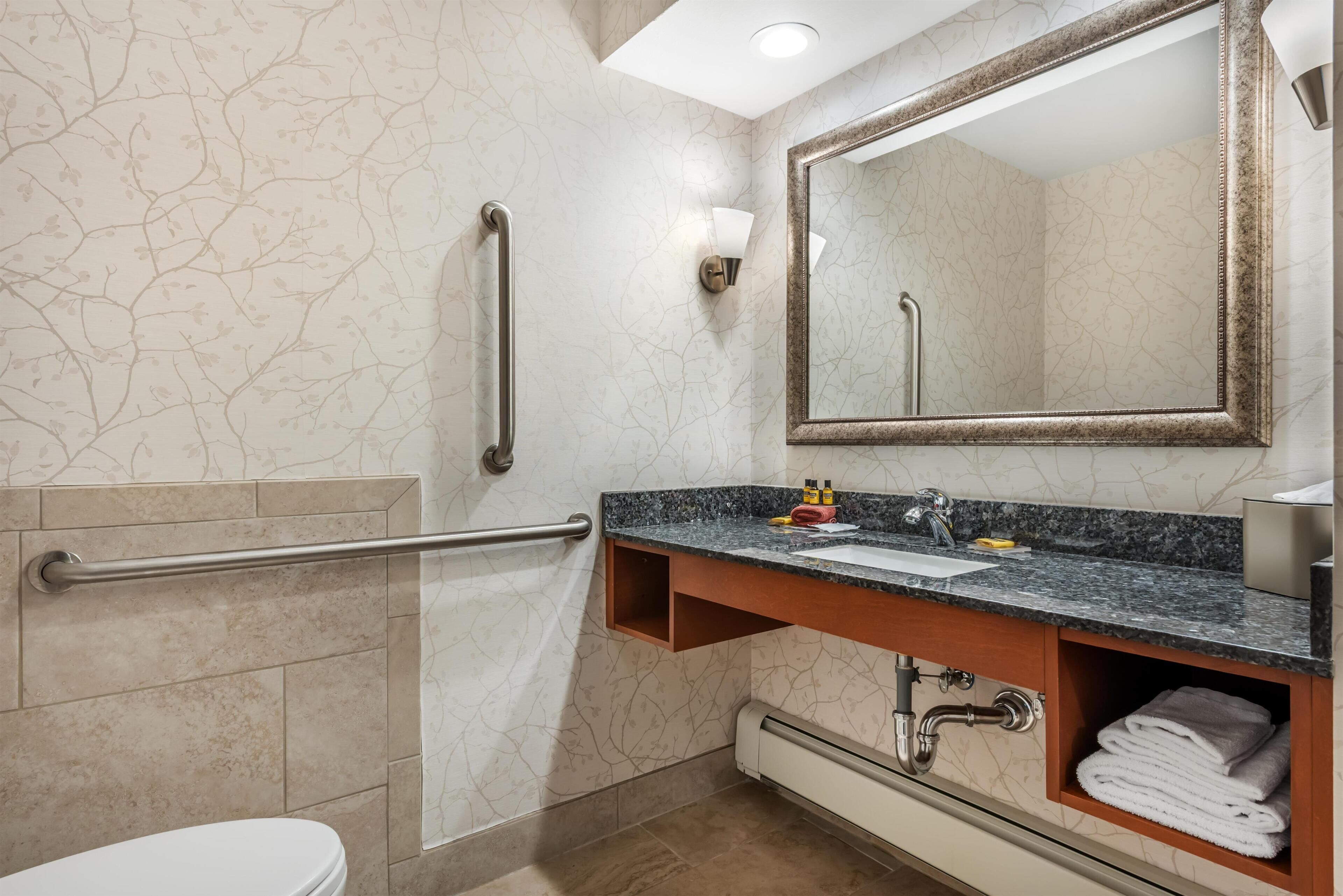 A bathroom with a marble wall, a sink with a mirror, a towel rack, and a toilet.