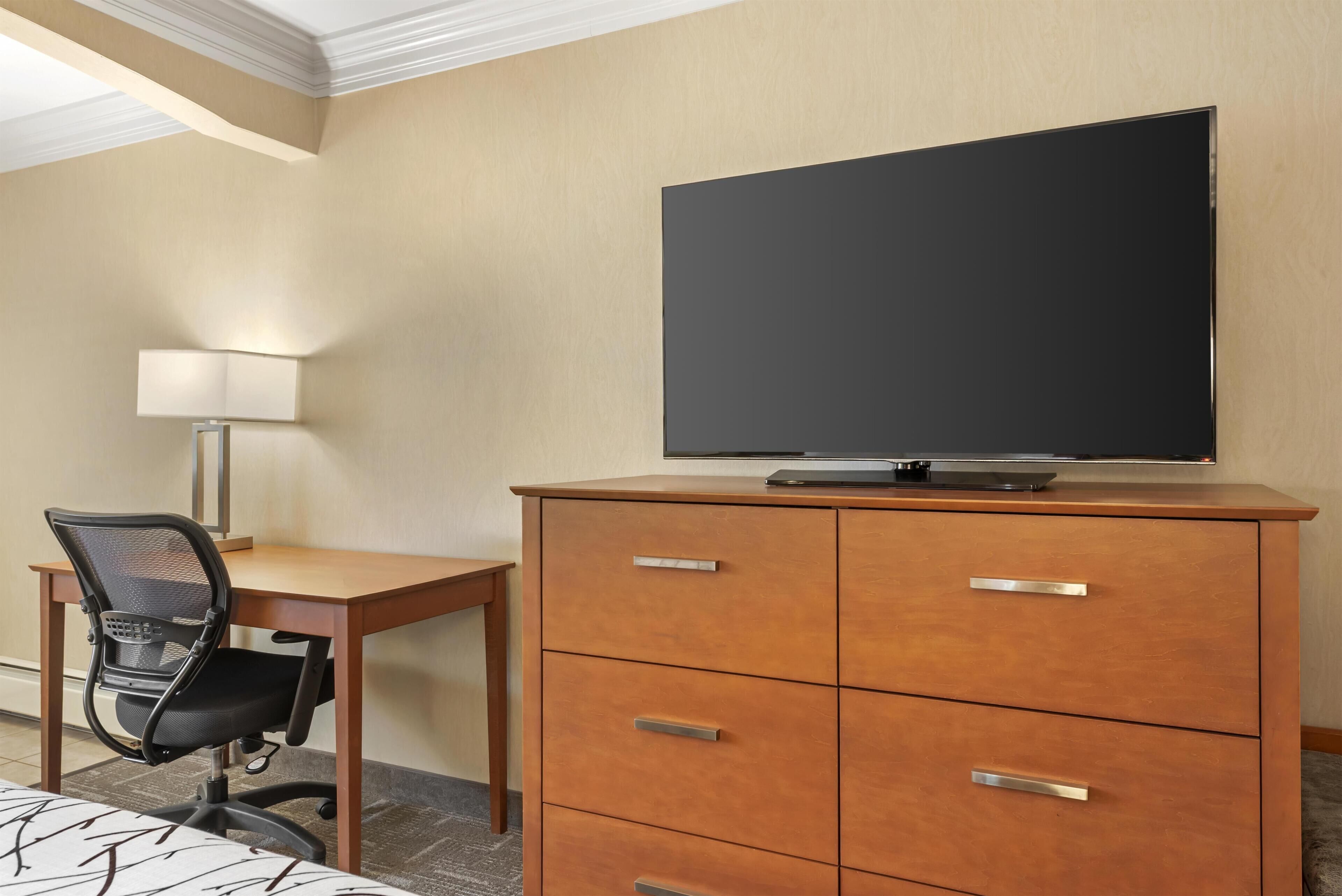 Een hotelkamer met een houten bureau, een zwarte bureaustoel, een flatscreen televisie op een houten kast en een lamp op het bureau.