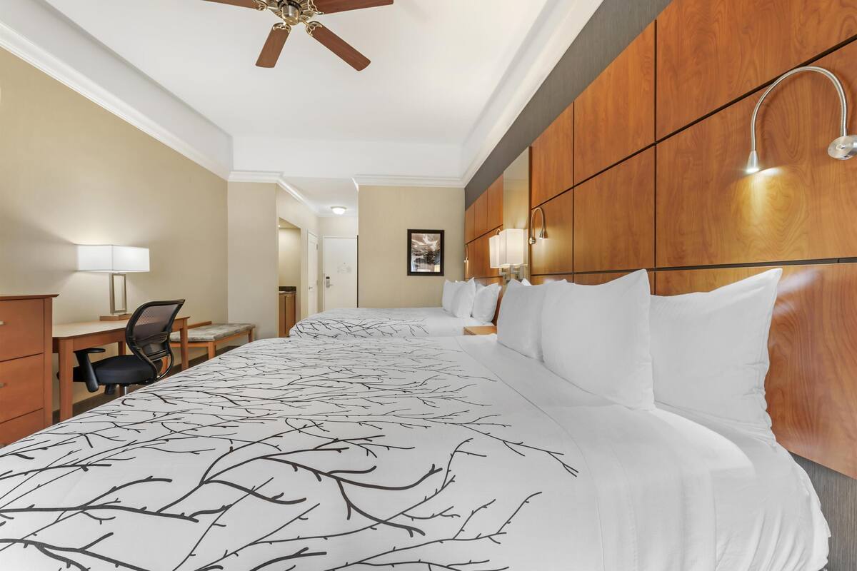 Une chambre d’hôtel avec un grand lit, un bureau, une chaise, un ventilateur de plafond et une tête de lit en bois agrémentée de lampes fixées au mur.