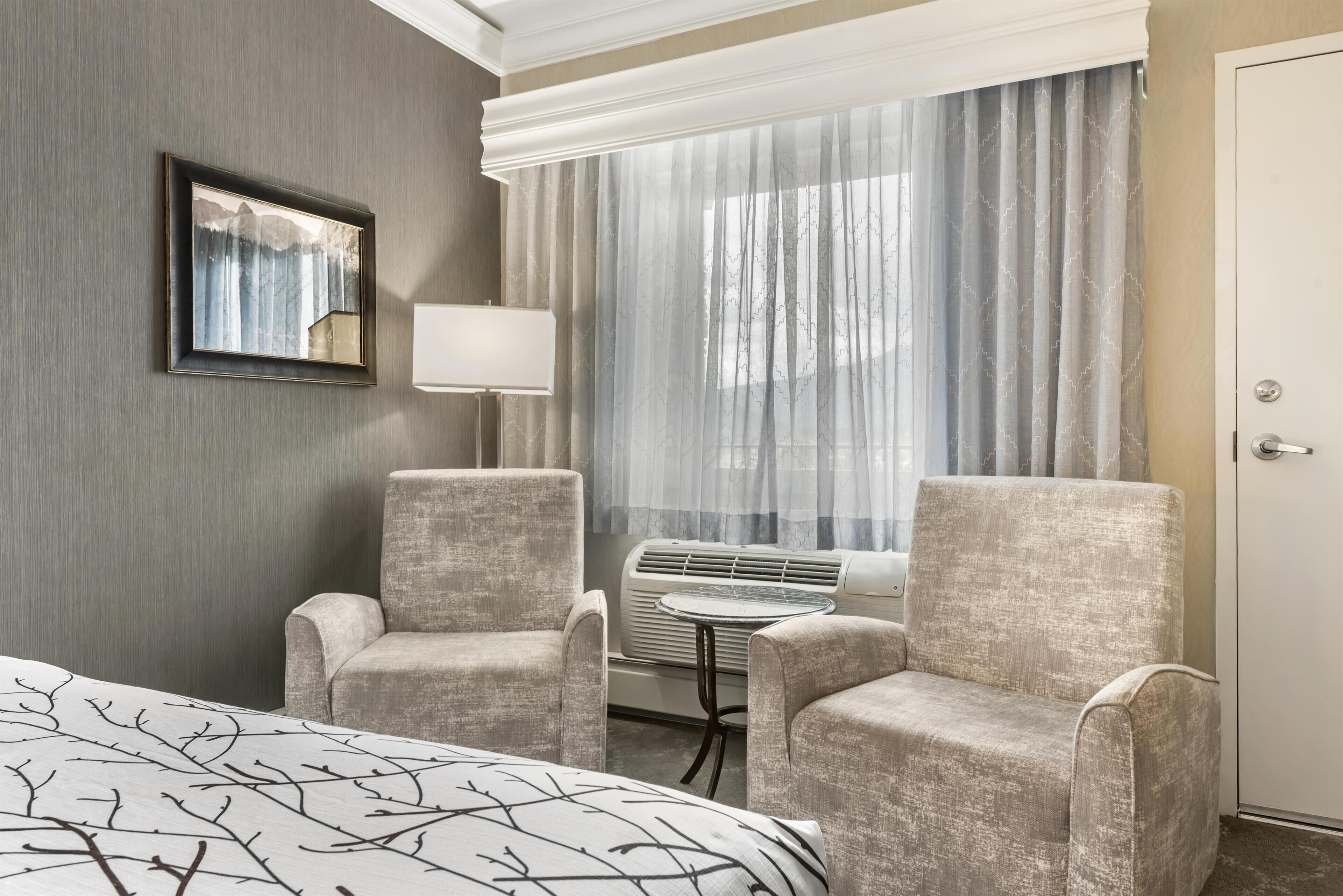 A hotel room with a bed, two armchairs, a table, a lamp, and a large window with curtains.