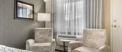 A hotel room with a bed, two armchairs, a table, a lamp, and a large window with curtains.
