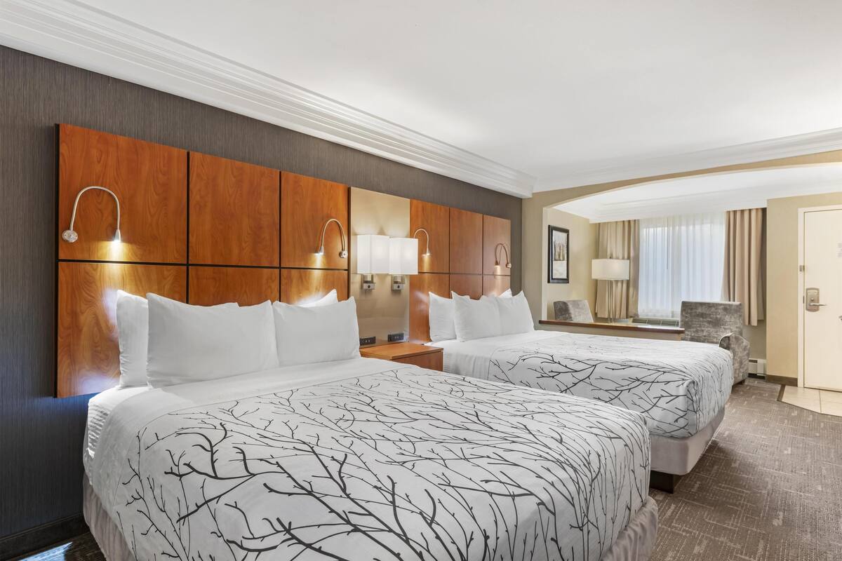 Une chambre d’hôtel avec deux lits, une tête de lit en bois, des lampes de chevet, un grand miroir et une vue sur le couloir.