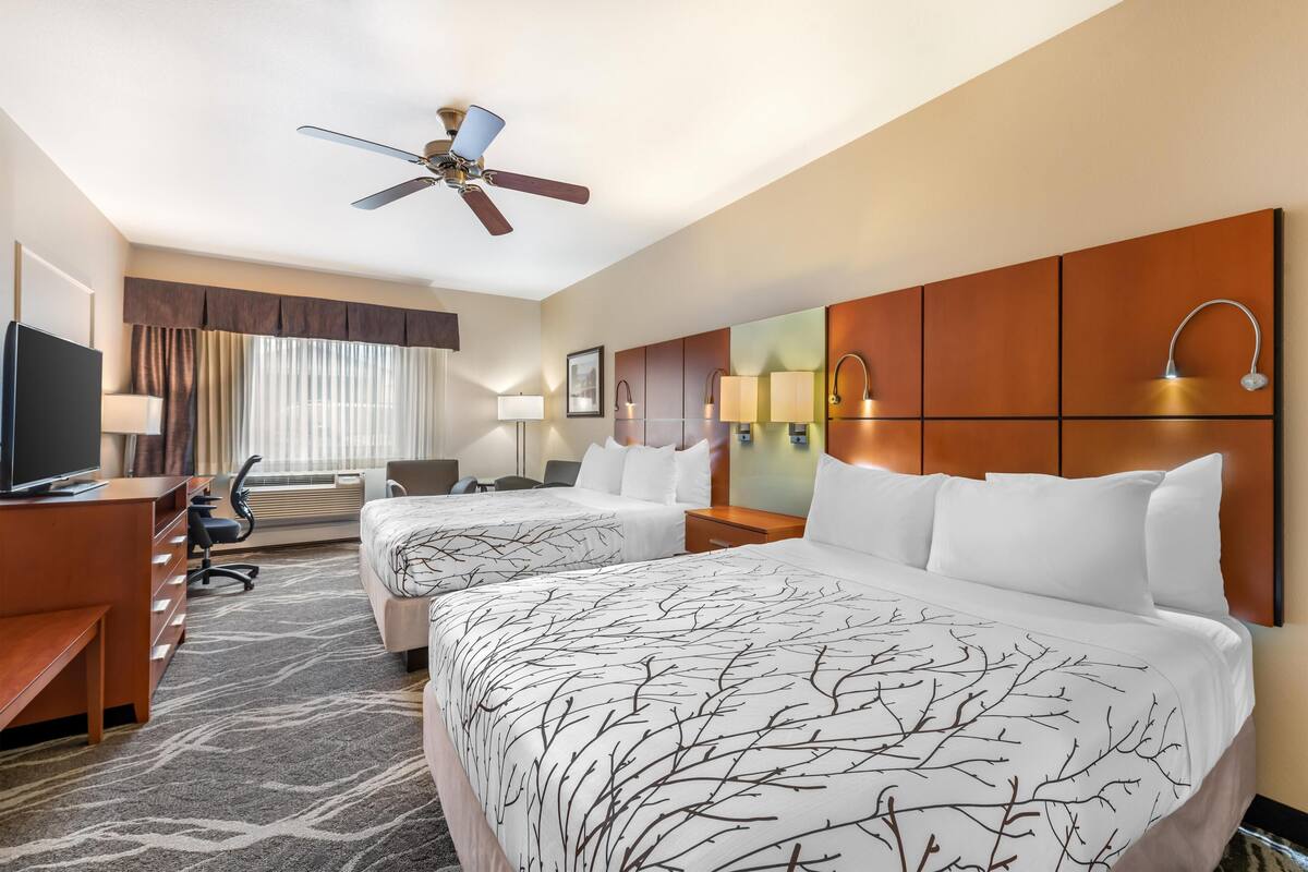 Une chambre d’hôtel avec deux lits, un bureau, une chaise, une télévision, un ventilateur de plafond et une grande fenêtre avec des rideaux.