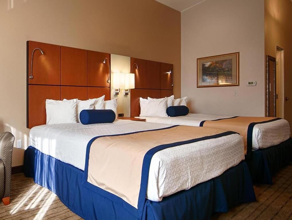 Habitació d'hotel amb dos llits, un capçal de llit de fusta, làmpades laterals, un quadre a la paret i un terra amb moqueta.