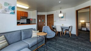 Ein modernes Hotelzimmer mit grauem Sofa, einem kleinen runden Tisch, einem Schreibtisch mit Stühlen, einer Küchenzeile und einem separaten Schlafbereich mit Bett und Nachttisch.