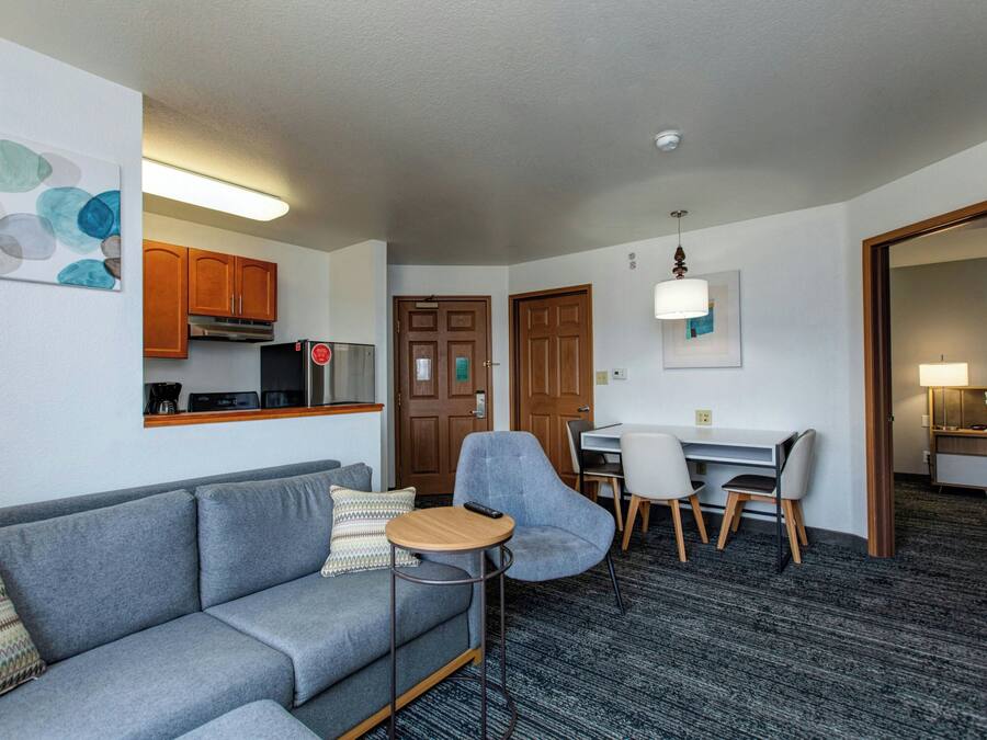 Une chambre d’hôtel moderne avec un canapé gris, une petite table ronde, un bureau avec des chaises, un coin cuisine et un espace nuit séparé comprenant un lit et une table de chevet.