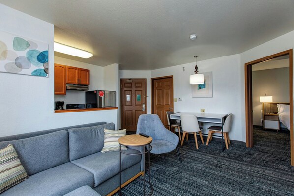 A modern hotel room with a grey sofa, a small round table, a desk with chairs, a kitchenette, and a separate sleeping area with a bed and nightstand. - Towneplace Suites By Marriott Denver Southwest (Littleton)
