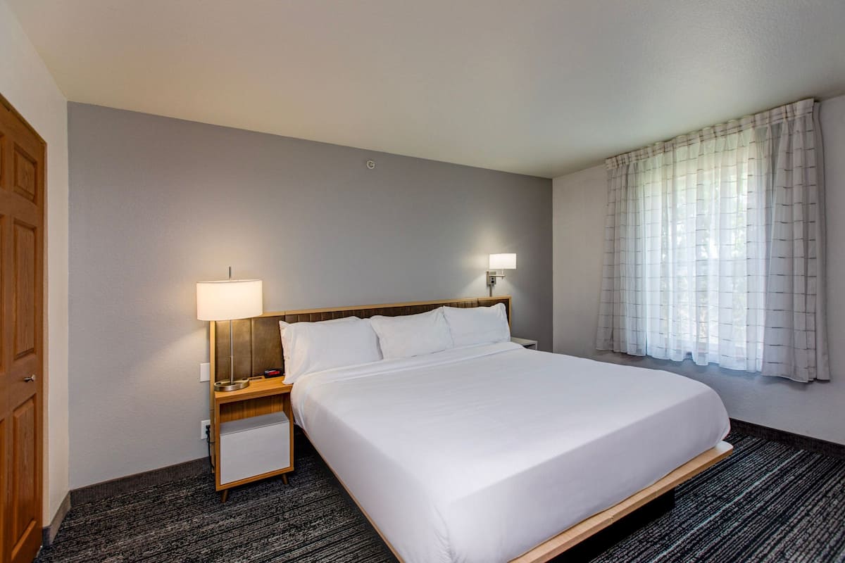 Ein Hotelzimmer mit einem großen Bett, zwei Nachttischlampen, einem Holznachttisch und einem Fenster mit durchsichtigen Vorhängen.