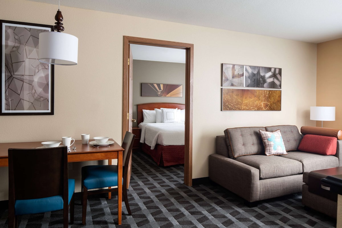 A hotel room with a bed, a sofa, a desk with chairs, and artwork on the walls.