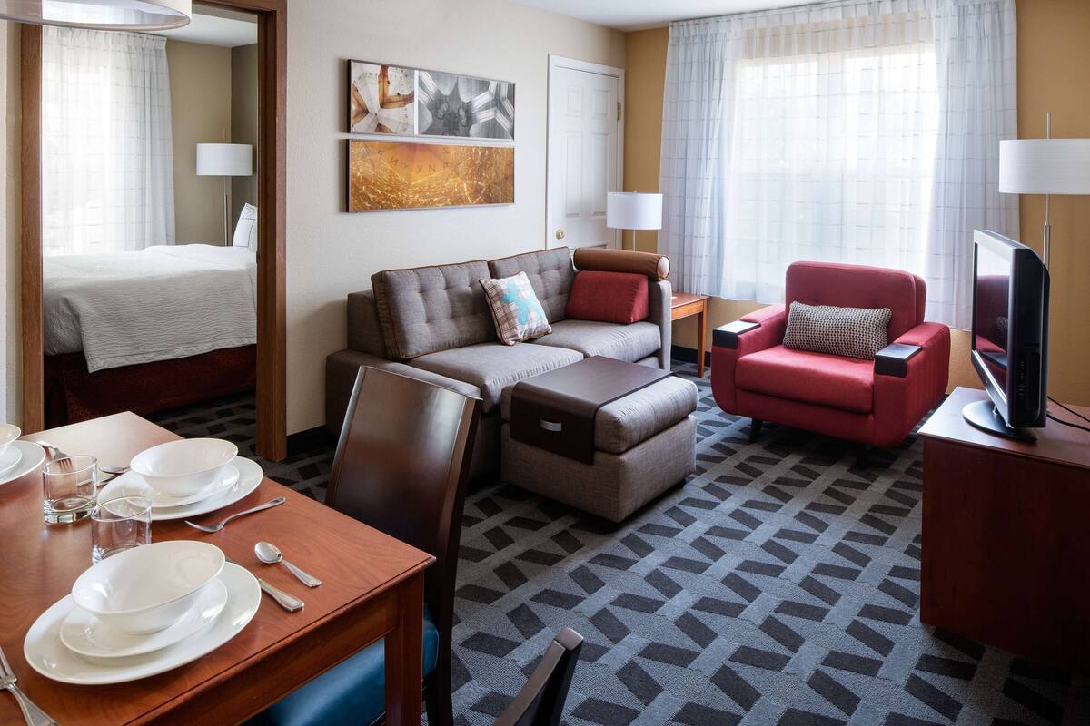 A hotel room with a bed, a sofa, a red armchair, a television, and a dining area with plates and glasses.