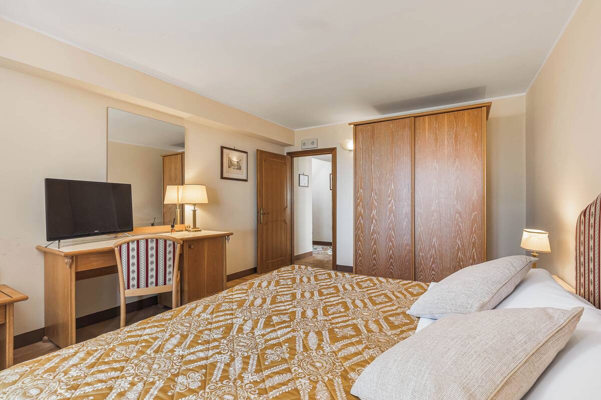 A hotel room with a bed, a desk with a television, a chair, a wardrobe, and a mirror.
