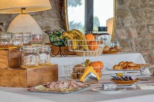 Un bufé de desayuno con diversos alimentos, como frutas, bollería y carnes, dispuesto sobre una mesa con una lámpara y una pared de piedra al fondo.