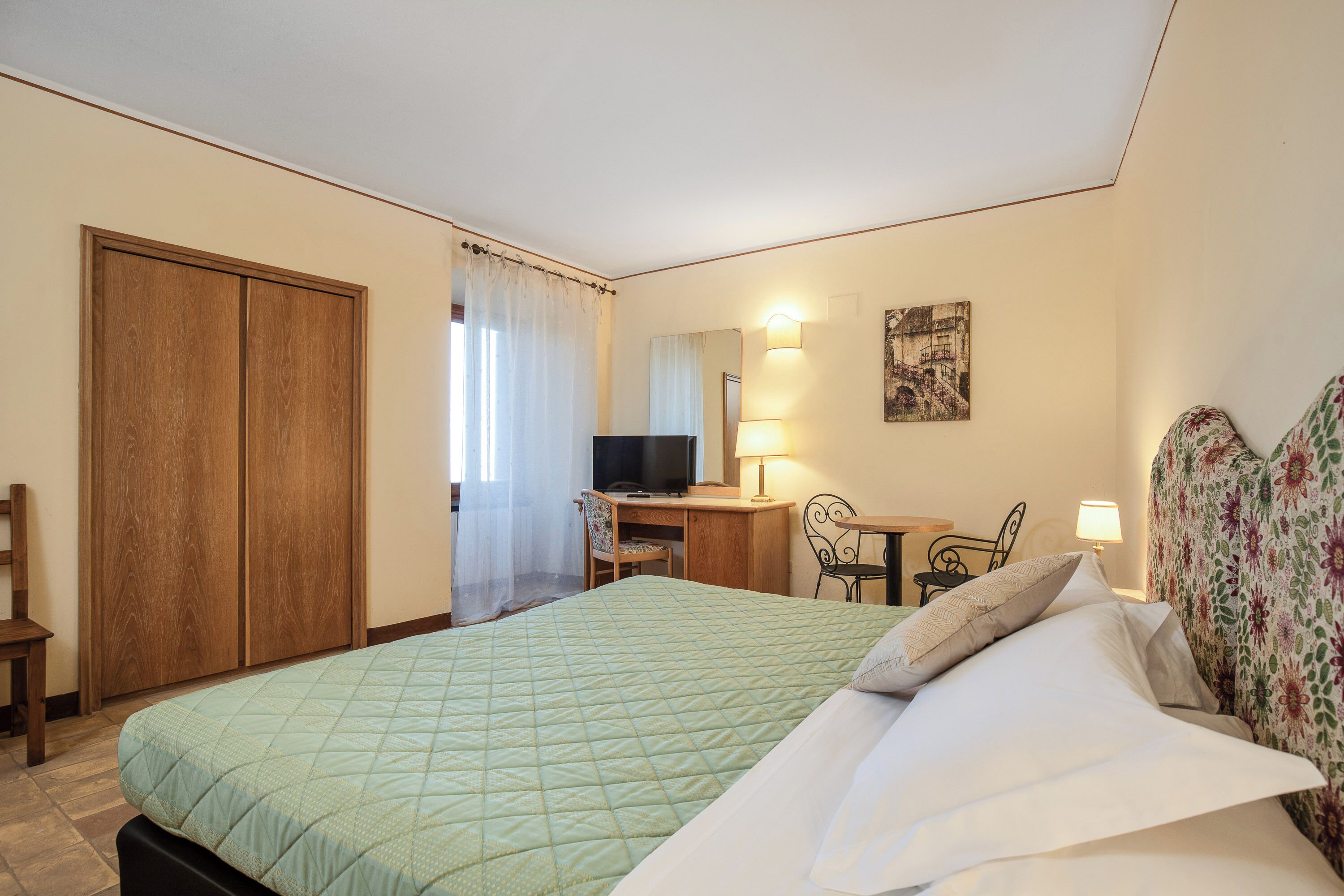 Una habitació d'hotel amb llit, escriptori, cadira, televisió i taula petita amb làmpada.