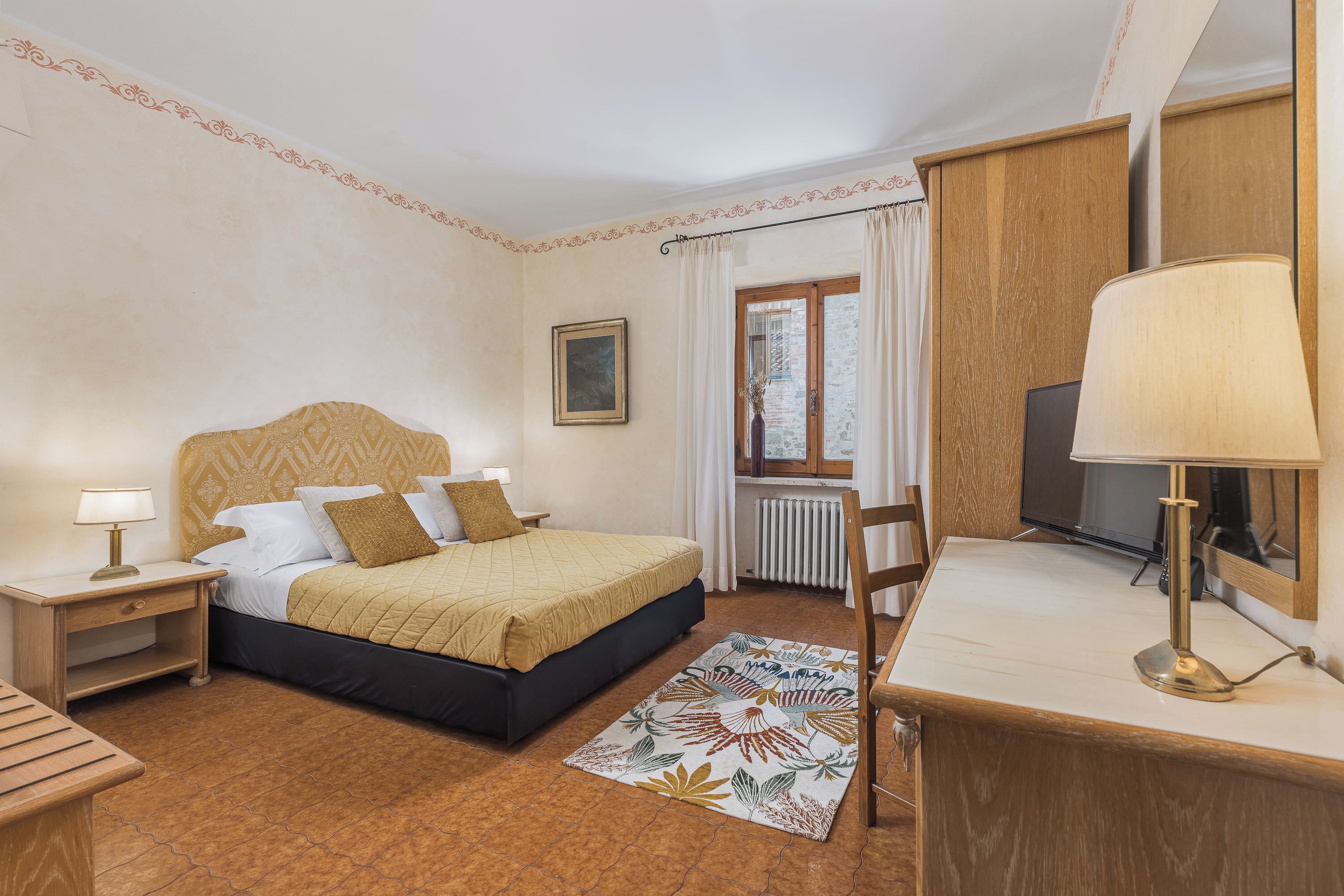 Ein Schlafzimmer mit Bett, Nachttischen, Schreibtisch, Stuhl, Fenster mit Vorhängen und Heizkörper.