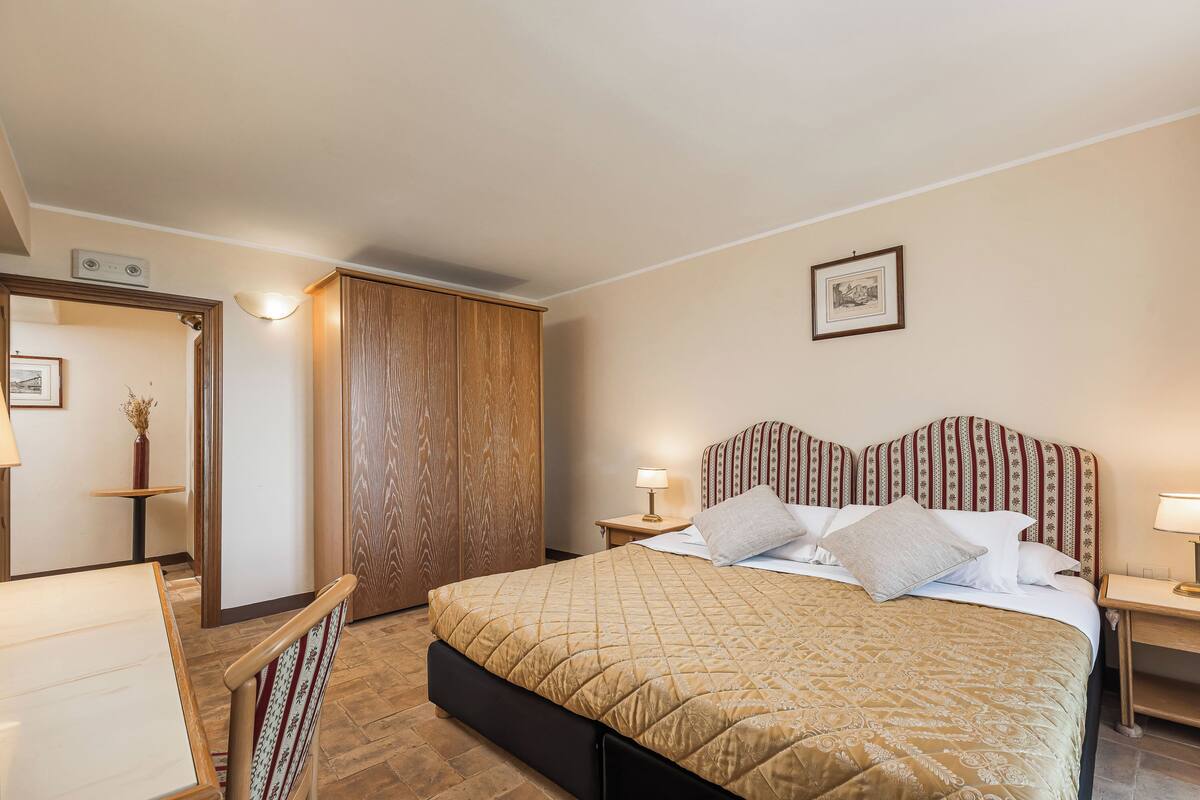 A hotel room with a large bed, a wooden wardrobe, a small table with a lamp, and a framed picture on the wall.