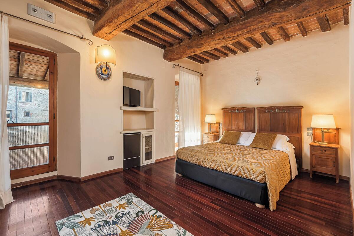 A bedroom with a wooden ceiling, a bed with a patterned bedspread, two bedside tables with lamps, a small cabinet, and a window with a view of a building outside.