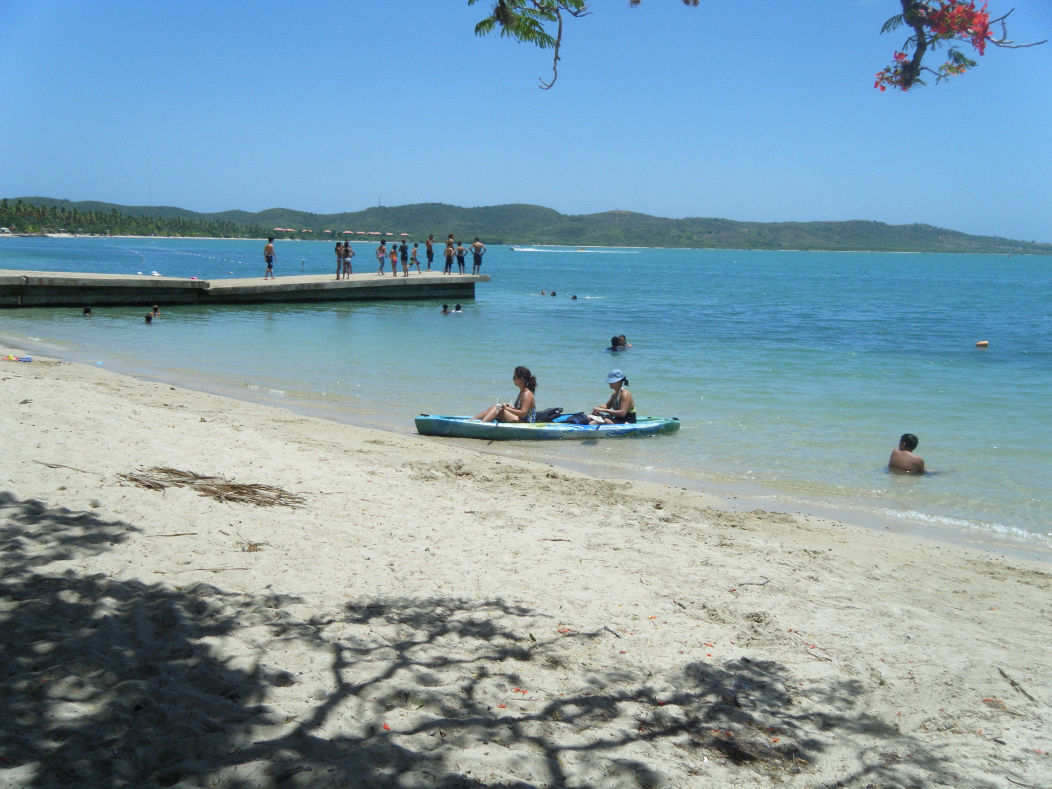 A beach with a dock, people in the water, and a small boat with two people.
