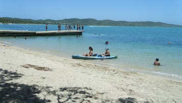 Una playa con un muelle, gente en el agua y un pequeño bote con dos personas.