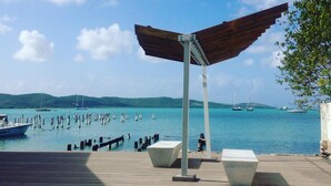A wooden deck with a metal structure, possibly a shelter, overlooking a calm body of water with boats and a distant shoreline. - Parador Boquemar (Cabo Rojo)