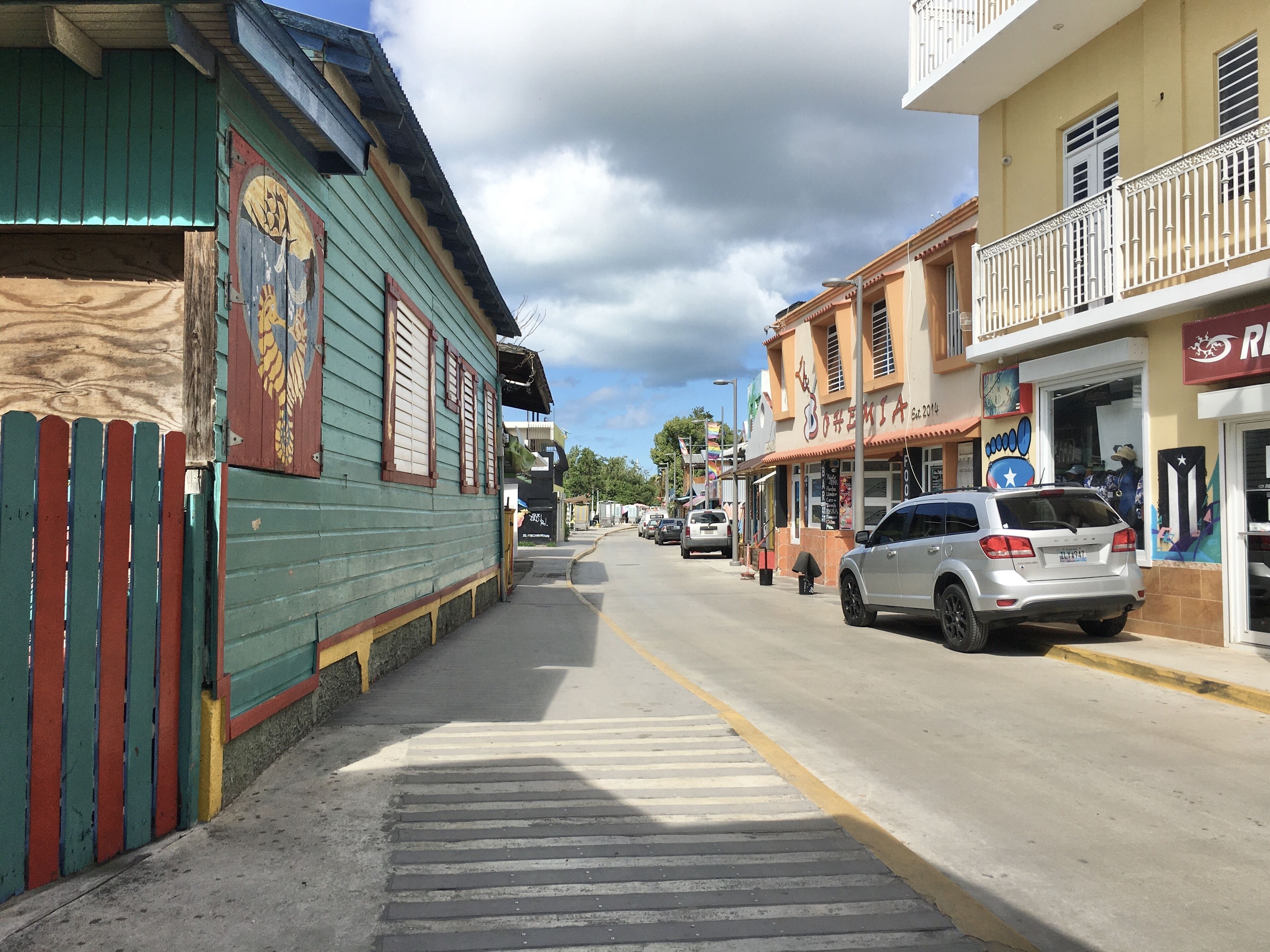Rua com edifícios, calçada e carros estacionados.