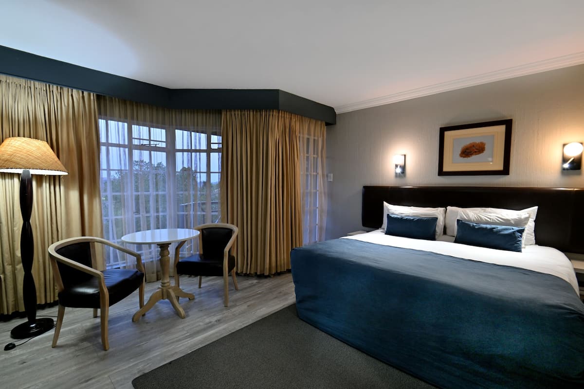 Une chambre d’hôtel comprenant un lit, deux chaises, une petite table, une lampe, une fenêtre avec des rideaux, un tableau encadré et un luminaire fixé au mur.