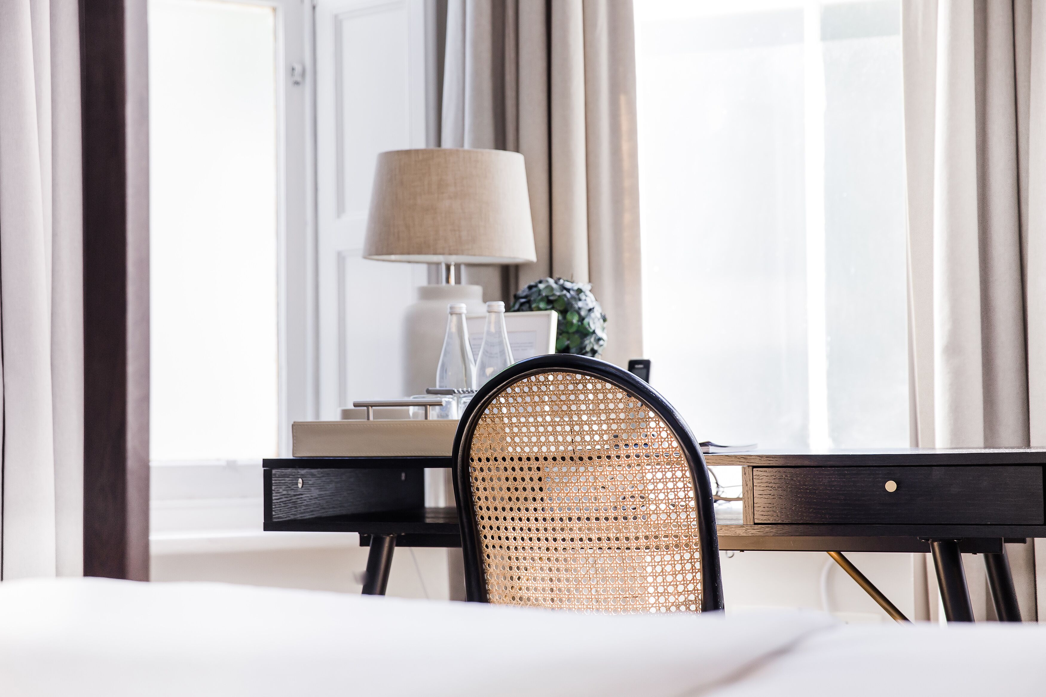 A hotel room with a bed, a desk with a lamp, a wicker chair, and a window with curtains.