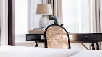 A hotel room with a bed, a desk with a lamp, a wicker chair, and a window with curtains.
